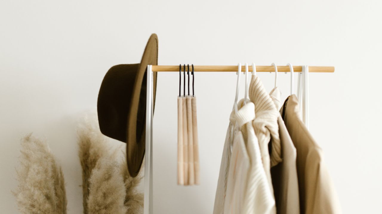 A mindful closet vibe emanates from the clothing rack adorned with neutral-toned clothes, a hat, and a hanging accessory against a pristine white wall. Pampas grass decor adds a serene touch nearby.